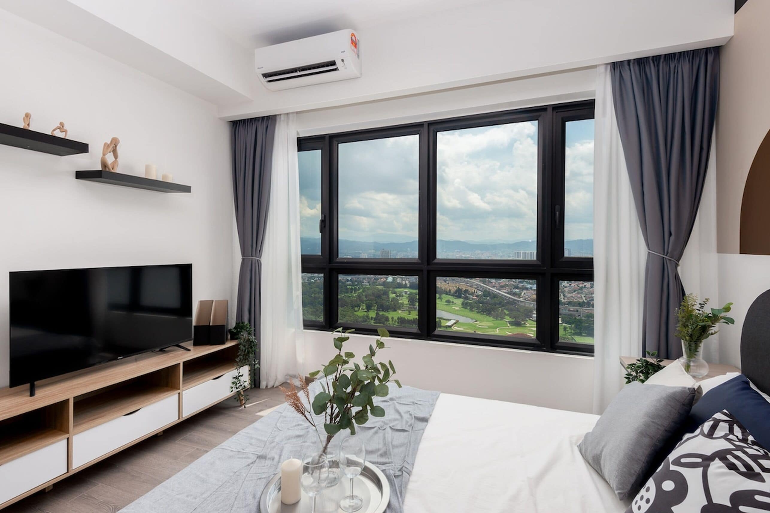A modern hotel room with a large window, a flat-screen TV, a bed with pillows, and a bedside table with a vase and glasses.