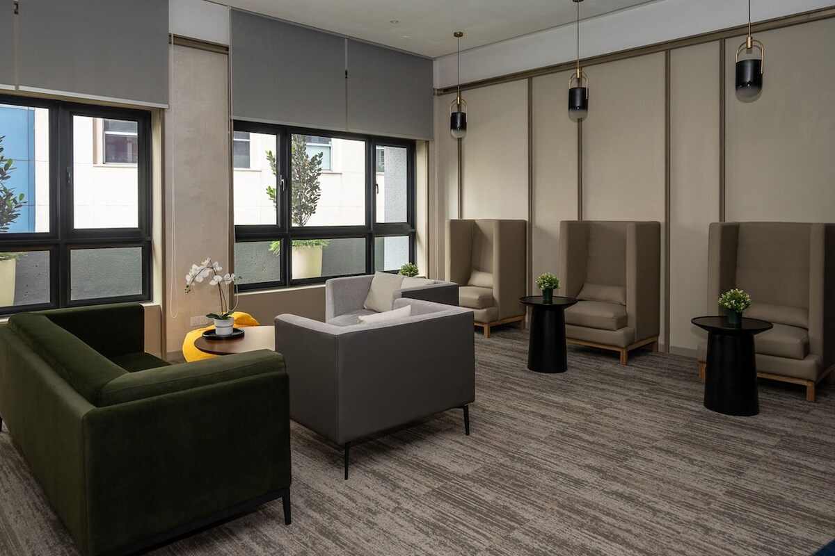Een moderne, goed verlichte kamer met grote ramen, een groene bank, een grijze fauteuil en twee kleine bijzettafels met potplanten.