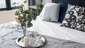 En snyggt bäddad säng med ett grått överkast, en dekorativ kudde med ett svartvitt blommönster och ett brickbord med ett ljus och två glas.