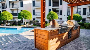 An outdoor barbecue area with a grill and a wooden cabinet, located near a swimming pool in a residential setting. - Beautiful 1Bd/1Bd Apartment in Dallas (Dallas)