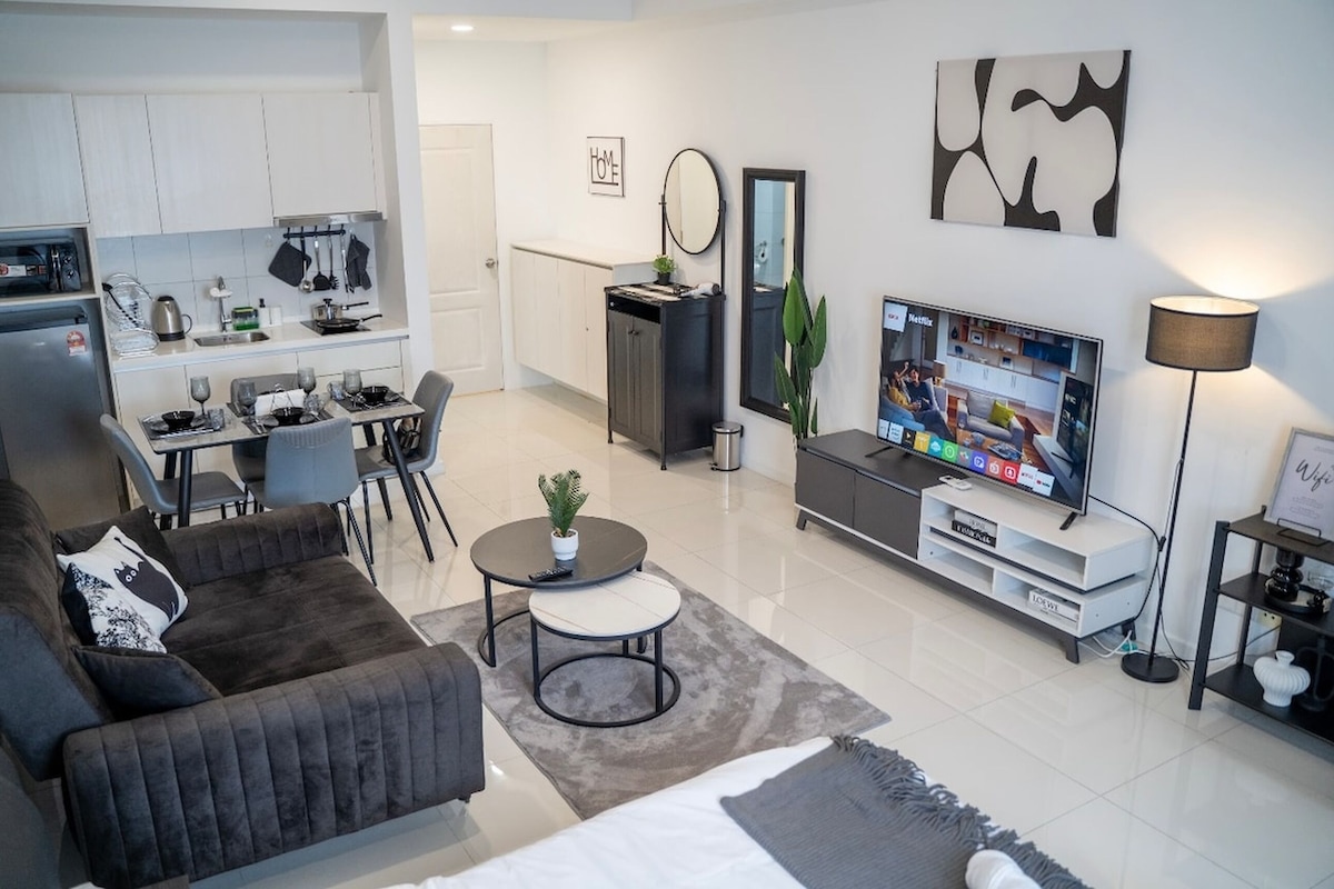 A modern living room with a sofa, a coffee table, and a TV. There is a kitchen area with a fridge, stove, and cabinets.