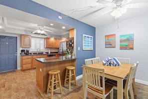 A modern kitchen with wooden cabinets, a central island with stools, a dining area with a wooden table and chairs, and artwork on the walls.