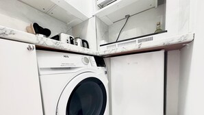 A compact laundry area with a front-loading washing machine, a small cabinet, and a countertop with a kettle and other items. - Studio Flat in Shepherd`s Bush, 2 minutes' walk away from Tube Station (London)