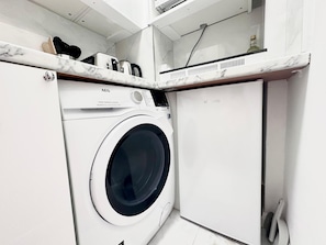 A compact laundry area with a front-loading washing machine, a small cabinet, and a countertop with a kettle and other items.