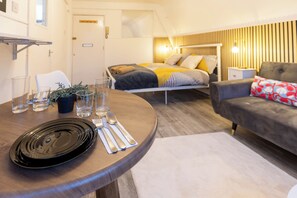 A modern room with a dining table set for two, a sofa, a bed with a headboard, and a small bedside table.