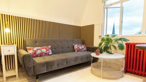 A modern living room with a grey sofa, a small white side table, a round white side table, a potted plant, and a red radiator. - Studio Flat in Shepherd`s Bush, 2 minutes' walk away from Tube Station (London)