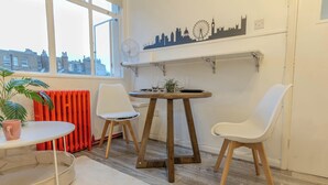 A room with a round table, two white chairs, a potted plant, a radiator, and a city skyline wall art. - Studio Flat in Shepherd`s Bush, 2 minutes' walk away from Tube Station (London)