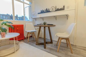 A room with a round table, two white chairs, a potted plant, a radiator, and a city skyline wall art.