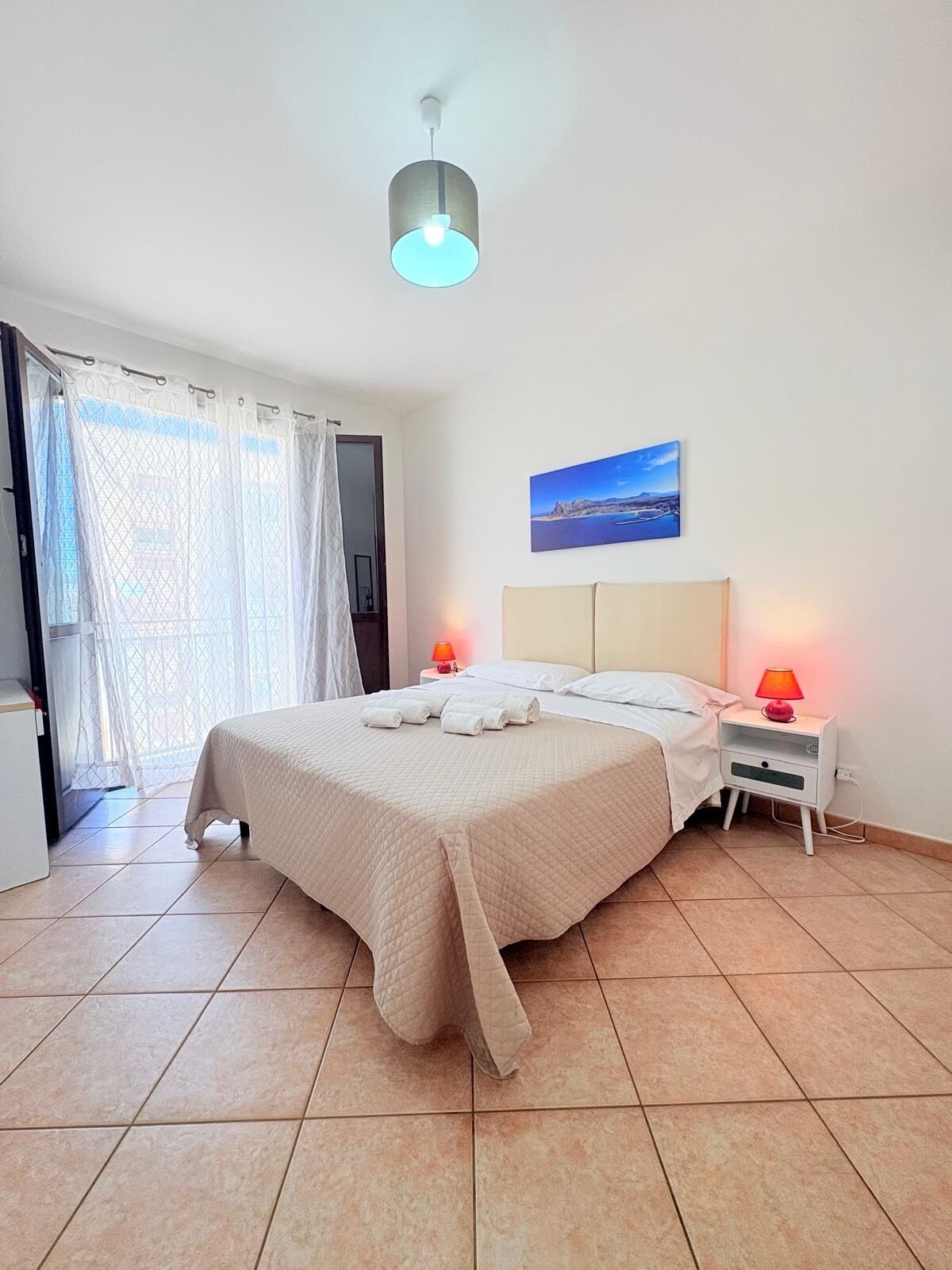Een slaapkamer met een bed, twee nachtkastjes met lampen, een raam met gordijnen en een schilderij aan de muur.