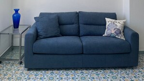 A modern living room with a blue sofa, a glass coffee table, a blue vase, a wall-mounted lamp, and a painting of a coastal scene. - Perla Bianca Holiday Home Ischia, Forio, nei pressi dei giardini Poseidon (Forio, Ischia (Na))