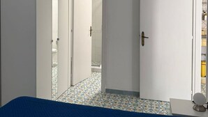 Ein Schlafzimmer mit blauer Bettdecke, einer Hängeleuchte und Blick in ein Badezimmer mit Fliesenboden.