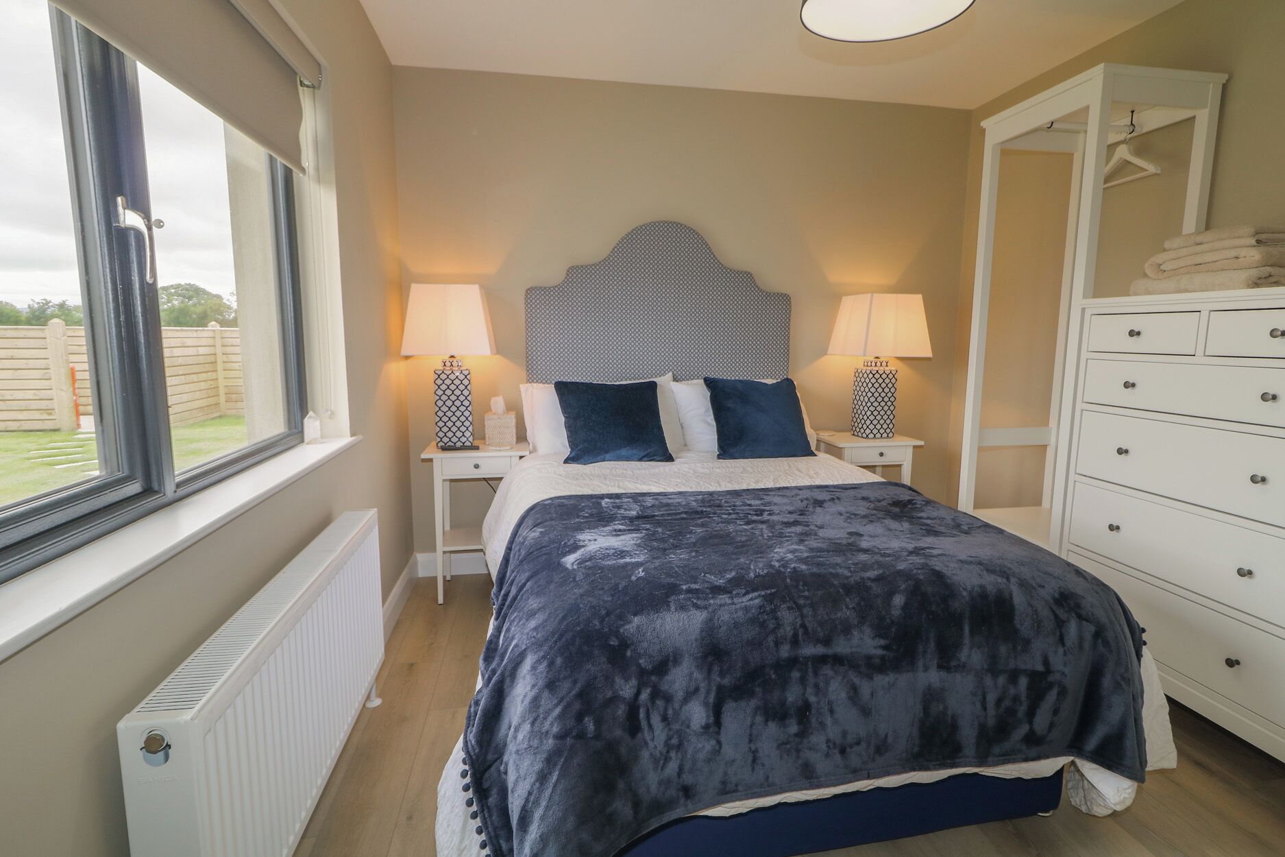 Un dormitorio con cama, dos mesitas de noche con lámparas, cómoda y ventana con vista a una valla.