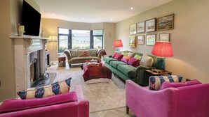 A living room with a fireplace, green and pink sofas, and a large window with a view of buildings. - Clarmes House (Limavady)