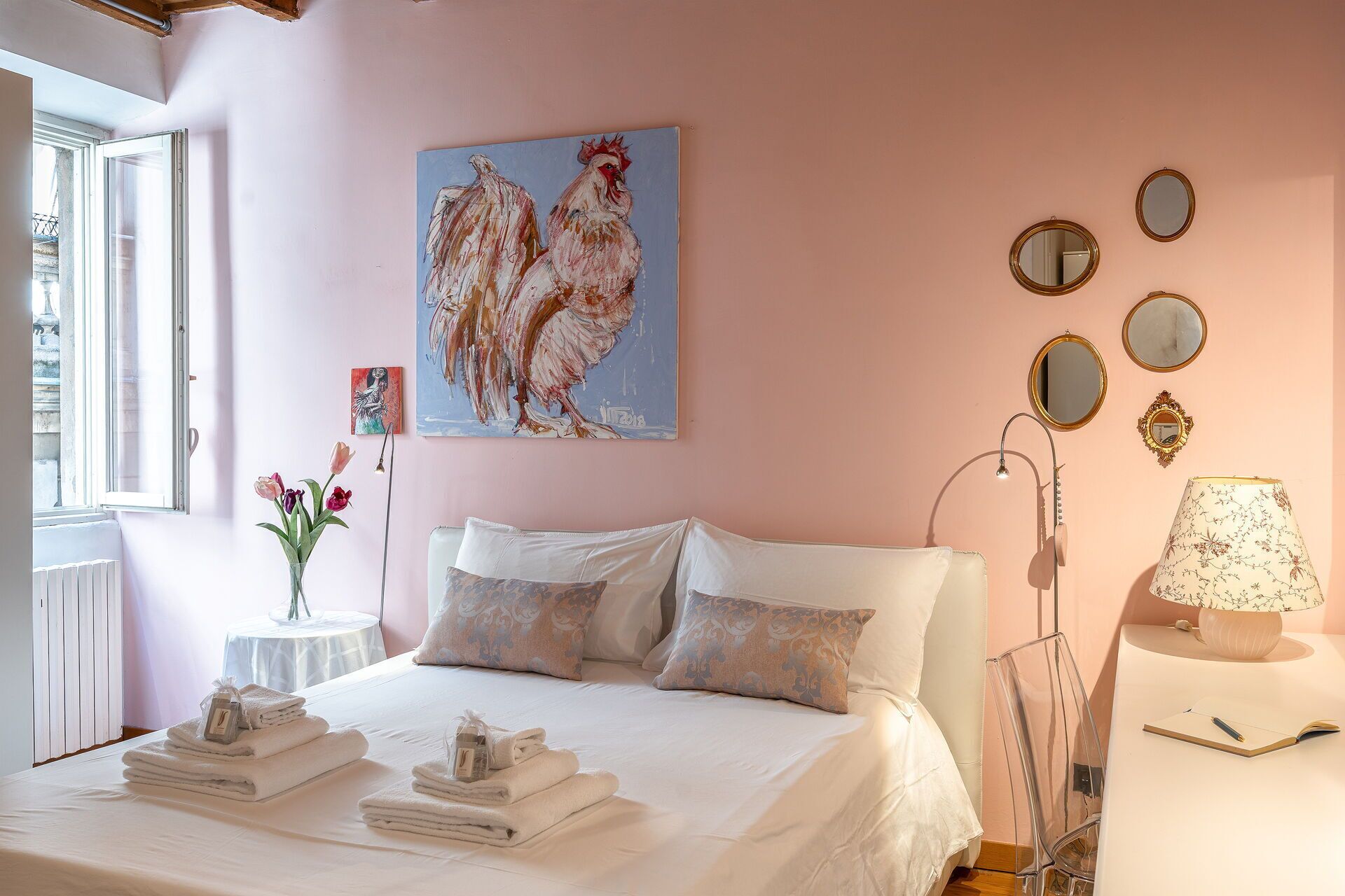 A bedroom with a bed, pillows, a painting, a vase with flowers, and a bedside table with a lamp.