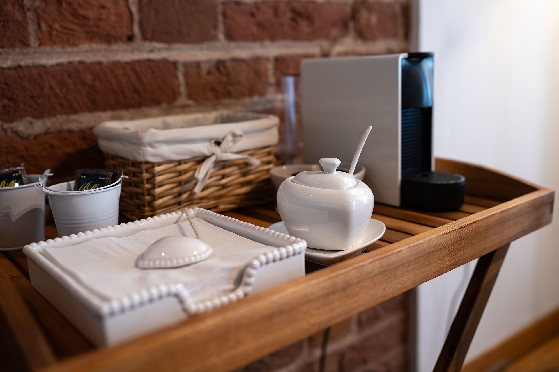 A wooden table with a white tray, a white sugar bowl, and a white sugar container. There are also two white mugs and a wicker basket in the background.