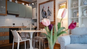 Une cuisine moderne avec des meubles gris, une table à manger blanche et des chaises. On peut voir un tableau représentant des poulets sur le mur, un vase avec des fleurs et un canapé blanc dans l’espace salon.