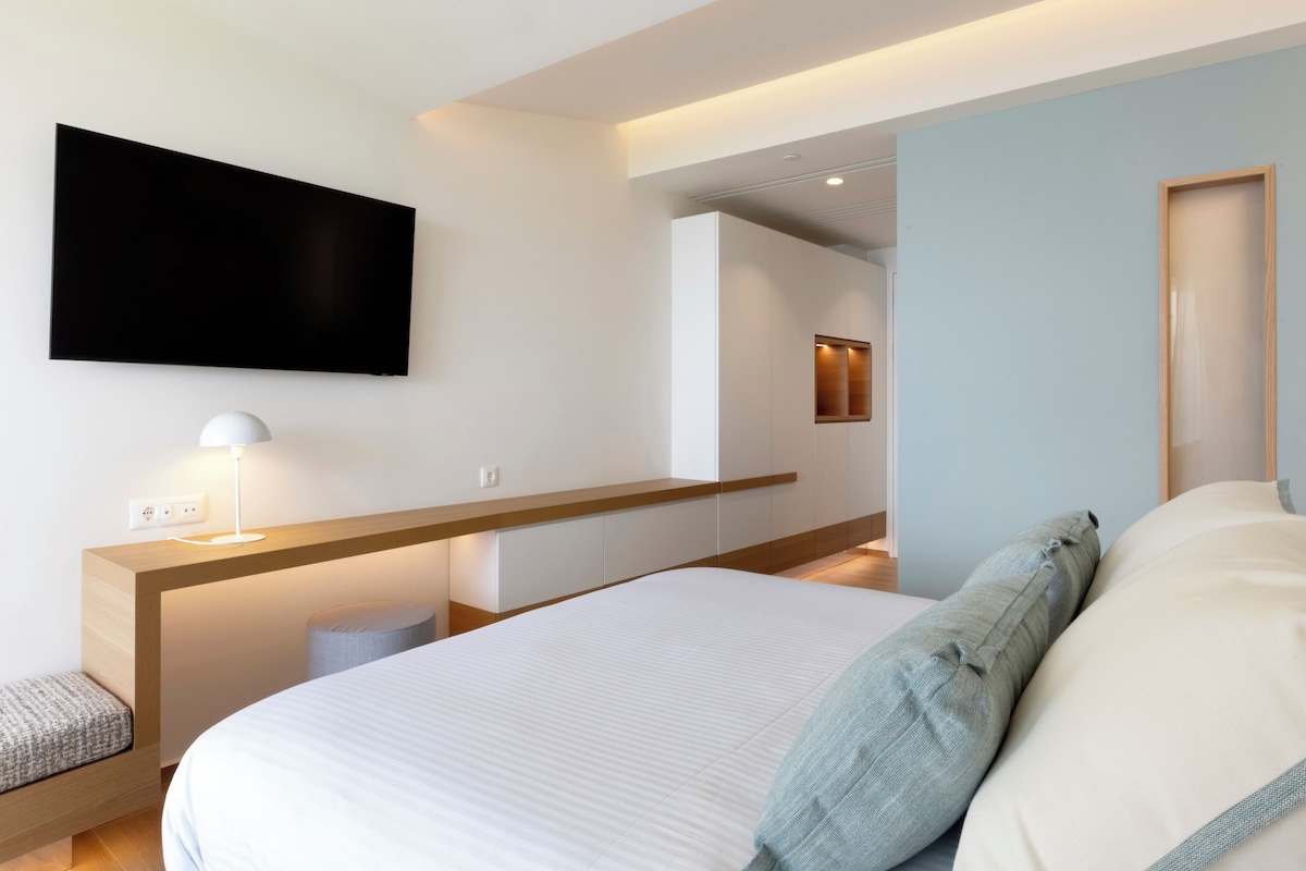 Una habitación de hotel moderna con una cama grande, una mesita de noche con lámpara, un televisor de pantalla plana en la pared y un cabecero de madera.