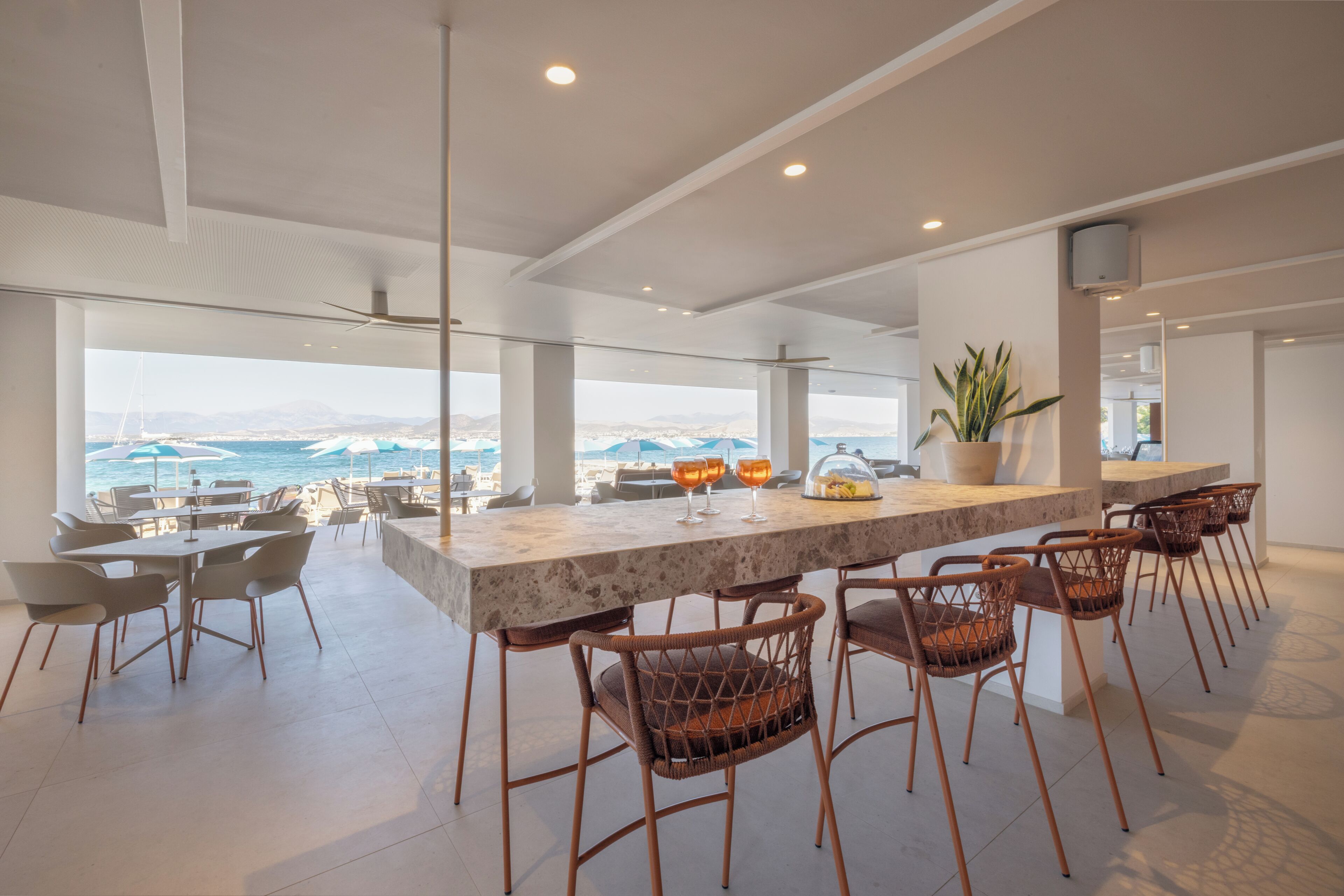 Zona de bar moderna con vista al mar, que cuenta con una encimera de mármol, sillas de mimbre y una planta en maceta.