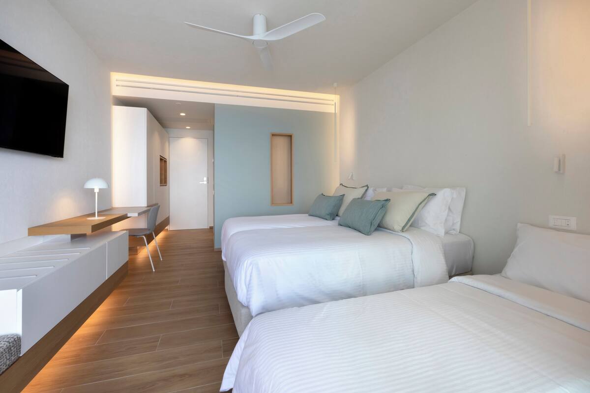 Ein Hotelzimmer mit zwei Betten, einem Schreibtisch, einem Sessel, einem Fernseher und einem Deckenventilator.