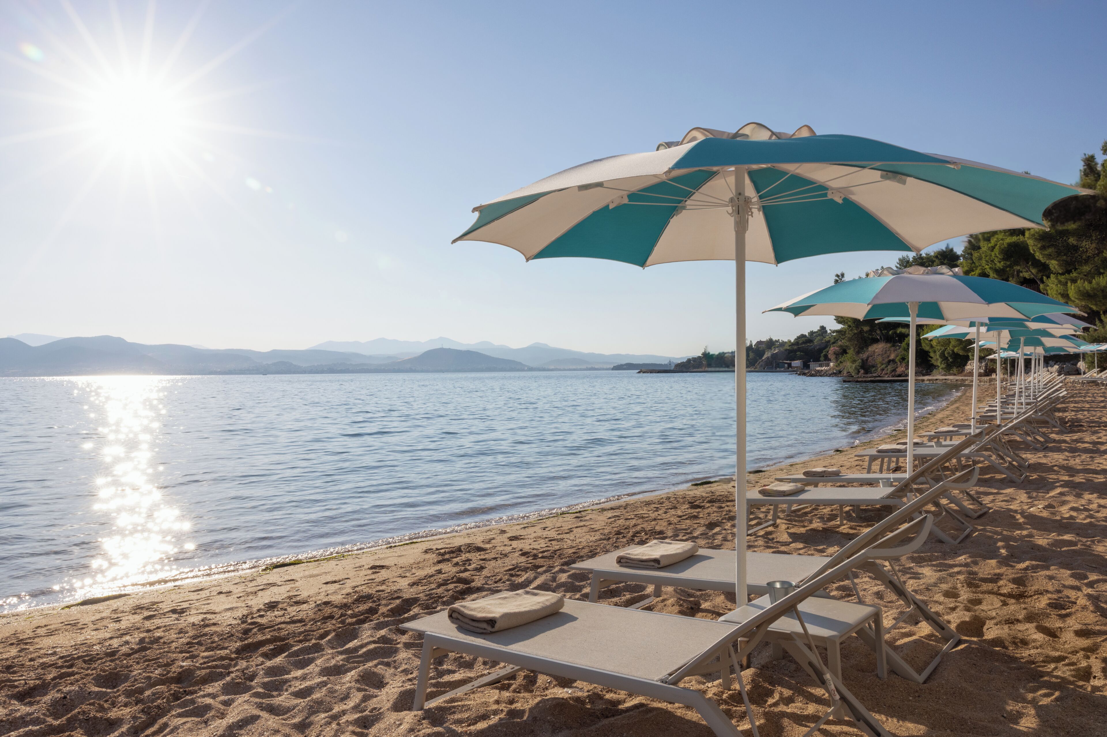 A beach with sun loungers and umbrellas, a calm sea, and distant mountains.