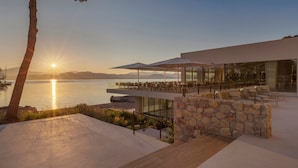 Un espace extérieur moderne, aménagé pour se détendre, avec vue sur le coucher de soleil sur l’eau, des murs en pierre et des rampes de protection en verre.