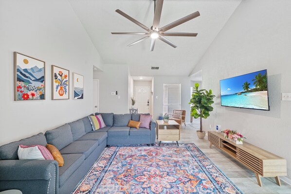 A living room with a grey sectional sofa, a patterned rug, a ceiling fan, and a TV displaying a beach scene.