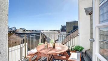 A round wooden table with four chairs, a bottle, and glasses on a balcony with a view of buildings.