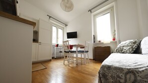 Habitació compacta amb llit, escriptori i cadira. Hi ha una televisió penjada a la paret, una finestra i una porta.