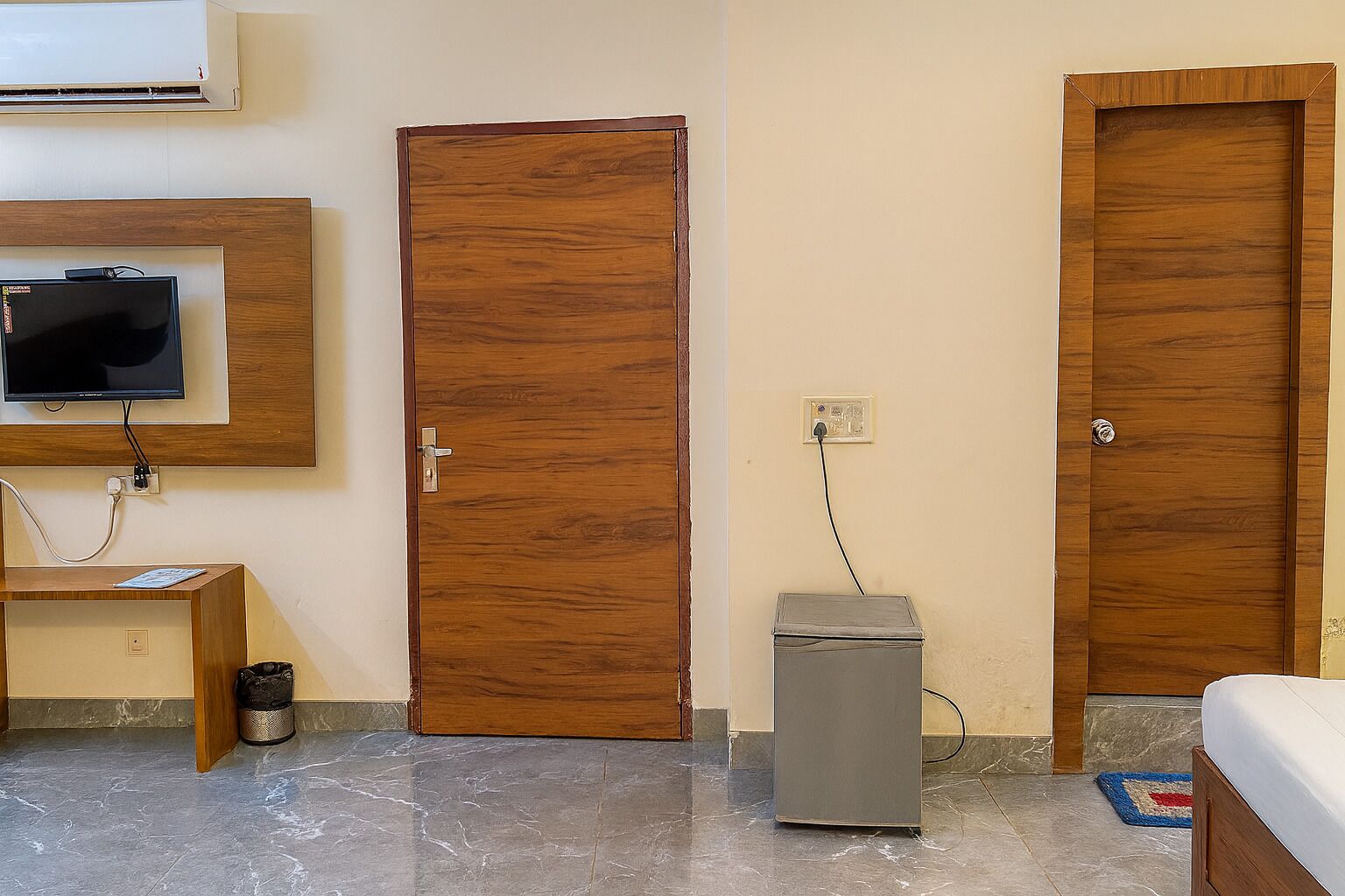 Une pièce avec une porte en bois, un petit meuble gris, un téléviseur fixé au mur et un lit recouvert d’un tapis bleu et rouge.