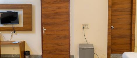 Une pièce avec une porte en bois, un petit meuble gris, un téléviseur fixé au mur et un lit recouvert d’un tapis bleu et rouge.