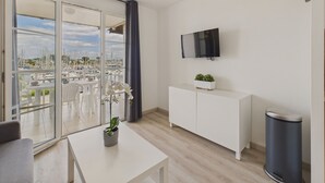 Un salon moderne doté d’une grande fenêtre, d’un téléviseur à écran plat fixé au mur, d’un meuble blanc et d’une petite poubelle.