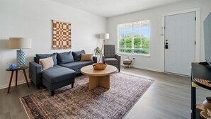 Una sala de estar con sofá, sillón, mesa de centro redonda y vista al exterior a través de una ventana.