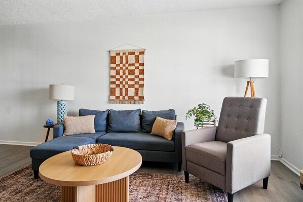 A modern living room with a grey sofa, a round wooden coffee table, a grey armchair, a patterned wall hanging, and a floor lamp.
