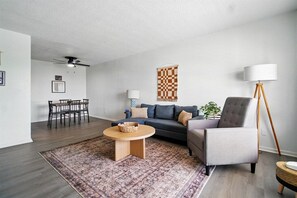 A living room with a sofa, armchair, coffee table, and a dining area with chairs and a table.