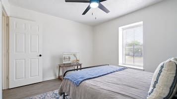 Une chambre à coucher avec un lit, un ventilateur de plafond, une fenêtre avec des stores, une porte et une pancarte de bienvenue.
