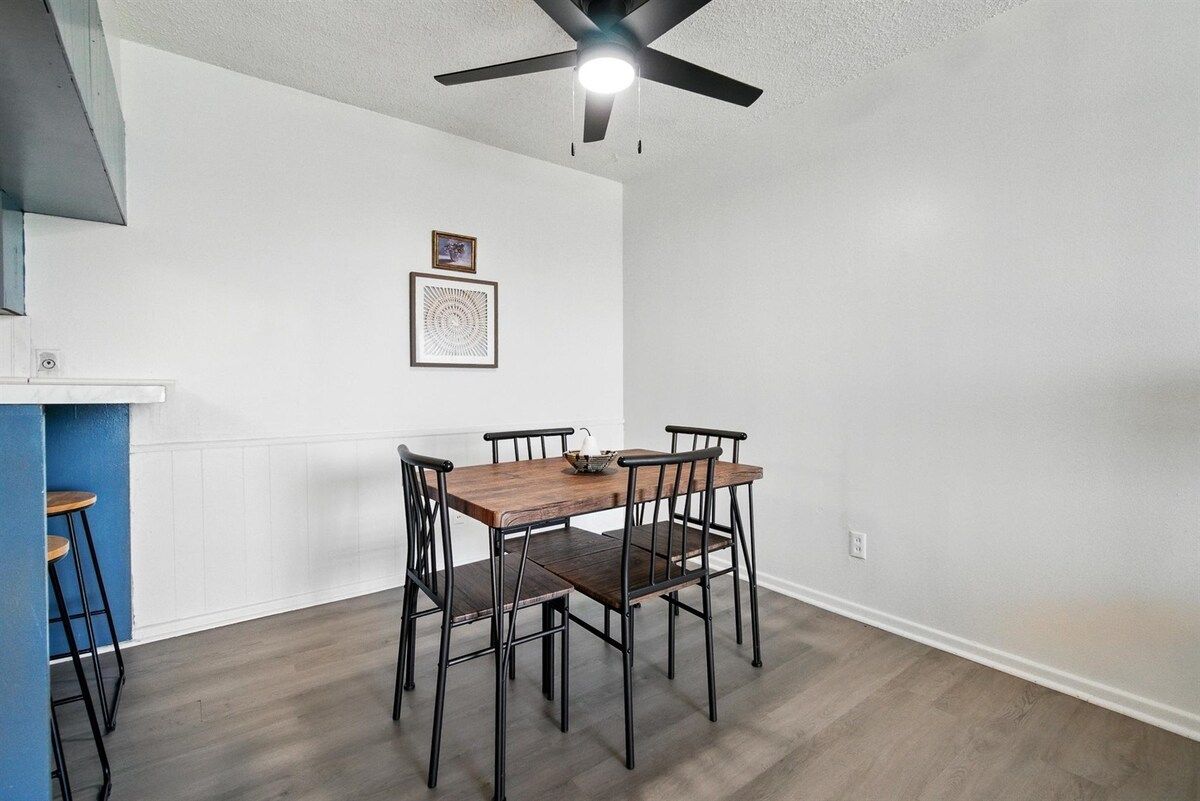 Un coin repas avec une table et des chaises en bois, un ventilateur de plafond et un mur blanc orné de deux tableaux encadrés.