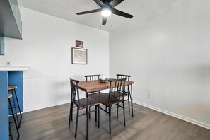 A dining area with a wooden table and chairs, a ceiling fan, and a white wall with two framed pictures.