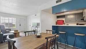 Una sala moderna con una mesa de comedor de madera, sofá gris y una cocina con armarios azules y un electrodoméstico rojo.