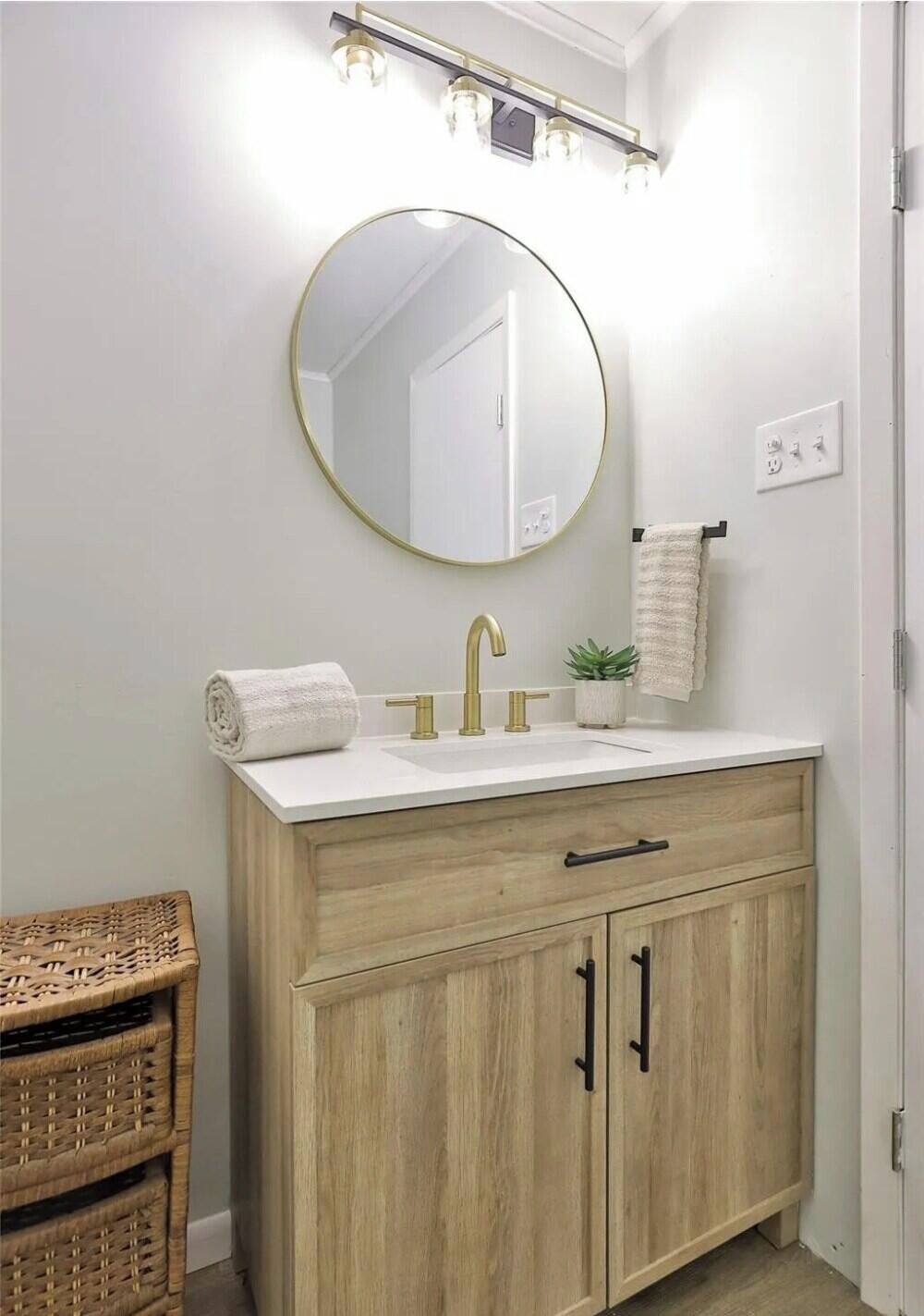 A bathroom with a round mirror, a sink with a gold faucet, wooden cabinets, a wicker stool, and a towel rack.