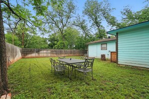 Un jardin avec une table et des chaises en métal, une clôture en bois et une maison d’un bleu clair.