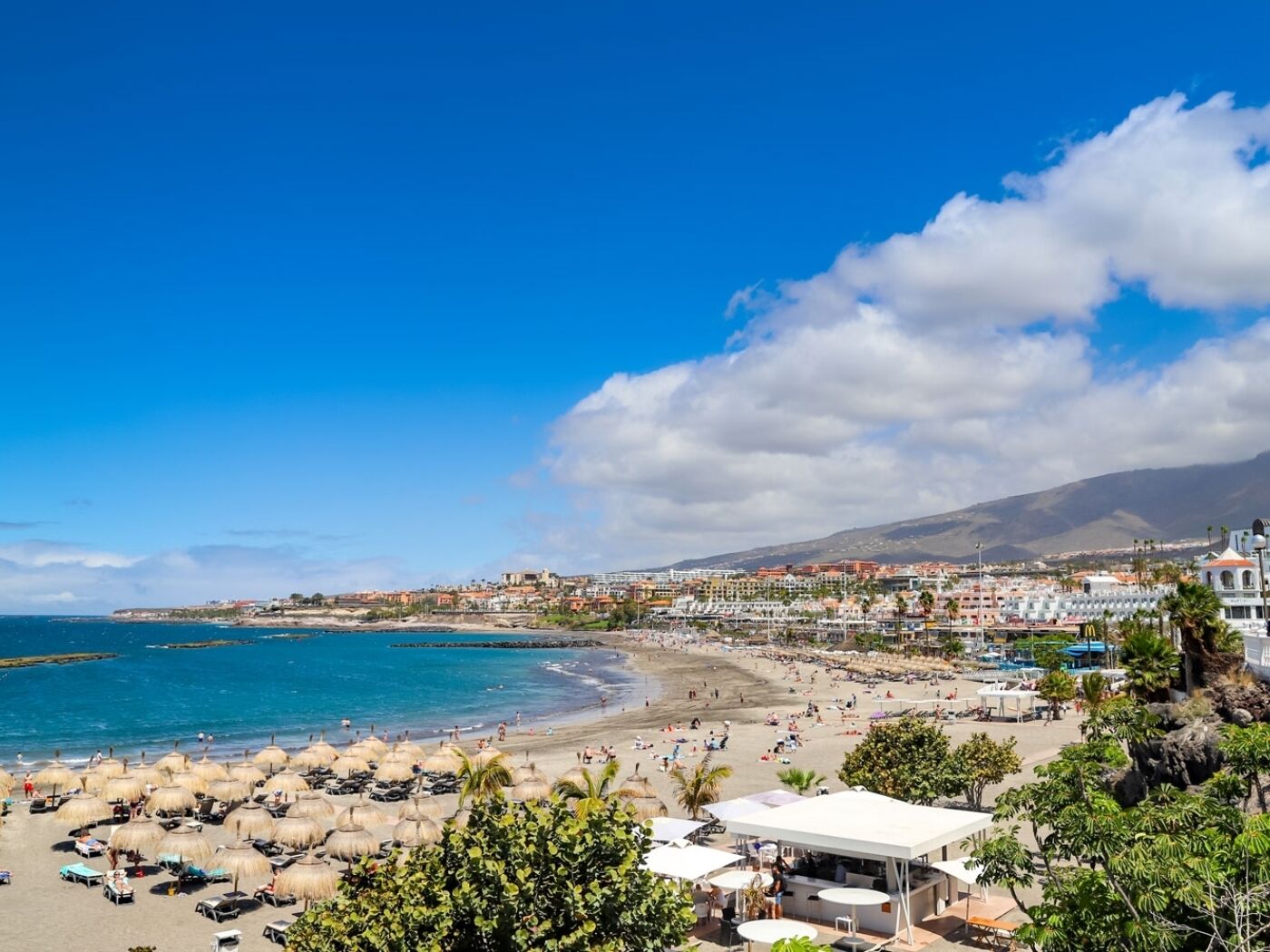 Una playa con sombrillas, un bar y vistas al océano y edificios al fondo.
