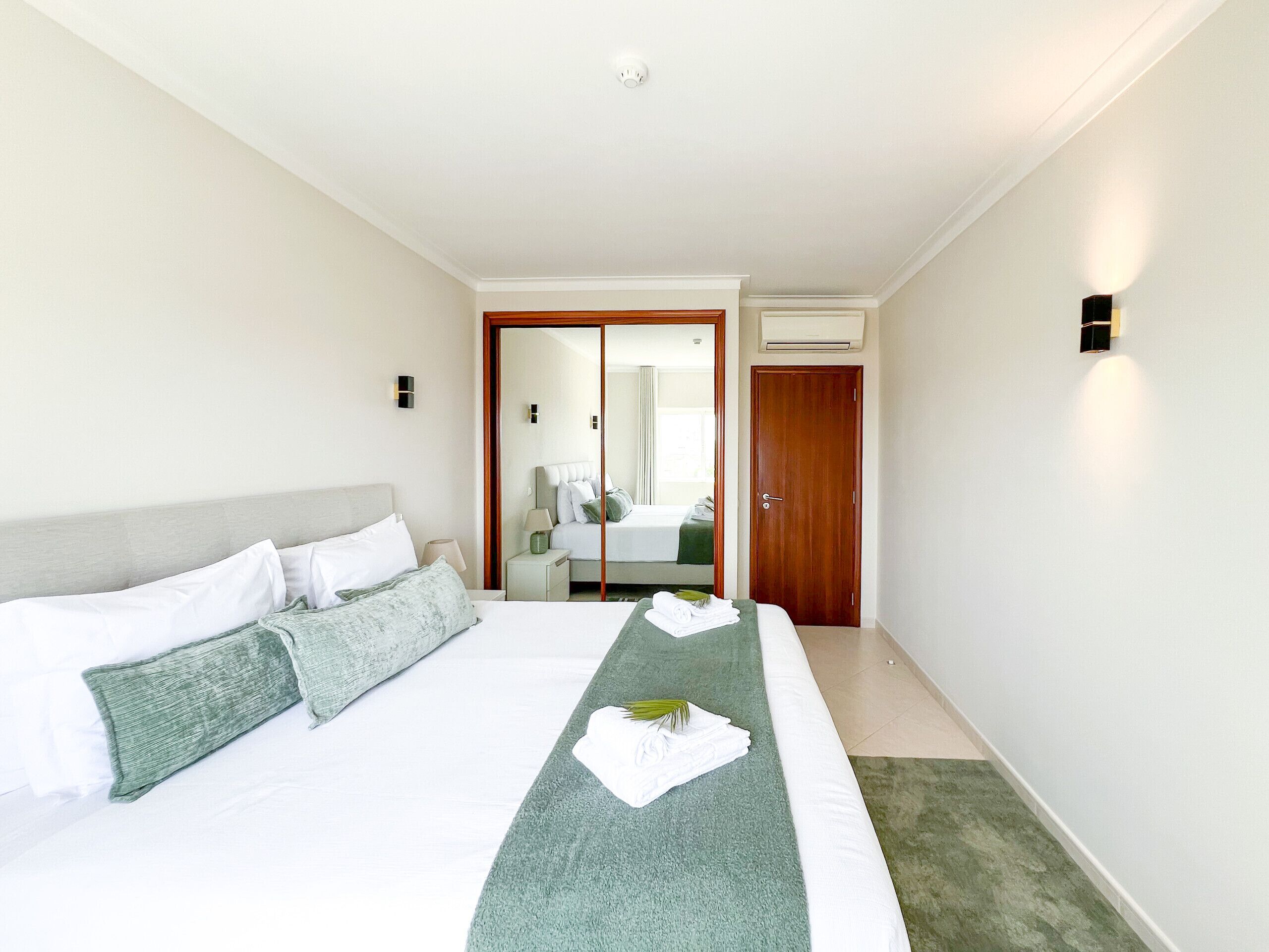 A hotel room with a large bed, a wooden door, and a mirror reflecting the room.