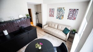 A modern kitchen with a black countertop, a herringbone backsplash, and a dining area with a black table and chairs. There is a sofa with a green pillow and abstract art on the wall. - Ralf Sannikov Apartment (3A) (Tartu)