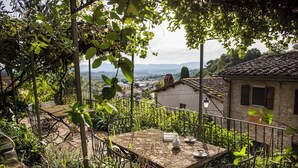 Un espacio de comedor al aire libre rústico con una mesa dispuesta para dos personas, rodeado de vegetación y con vistas a un pueblo de edificaciones tradicionales de piedra.