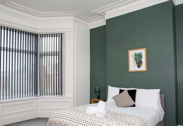A hotel room with a bed, a window with blinds, a nightstand with a lamp, and a framed picture on the wall.