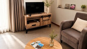 Un salon avec un téléviseur posé sur un meuble en bois, un canapé, une table basse avec des magazines et une plante.