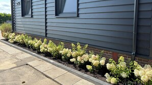 A house with a grey siding exterior, a brick foundation, and a flower bed with white flowers along the front. - Luxury Hay Barn Holiday Home with Open Countryside Views in the Green Heart (Alphen aan den Rijn)