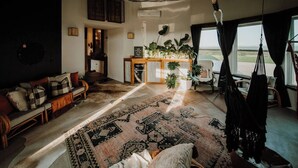 Una sala de estar acogedora con techo rústico, un sofá con cojines estampados, una mesa de centro con plantas y vistas al exterior a través de amplios ventanales.