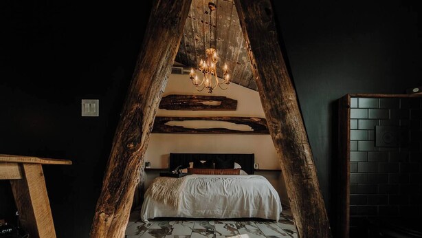 Una habitación con techo inclinado, una cama con cubrecama blanco, una araña y una pared oscura con textura.