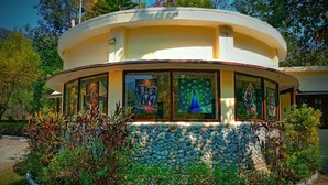 A circular building with a stone base, surrounded by a garden with various plants and a peacock mural. - The Harikanta Corbett Resort (Marchula)